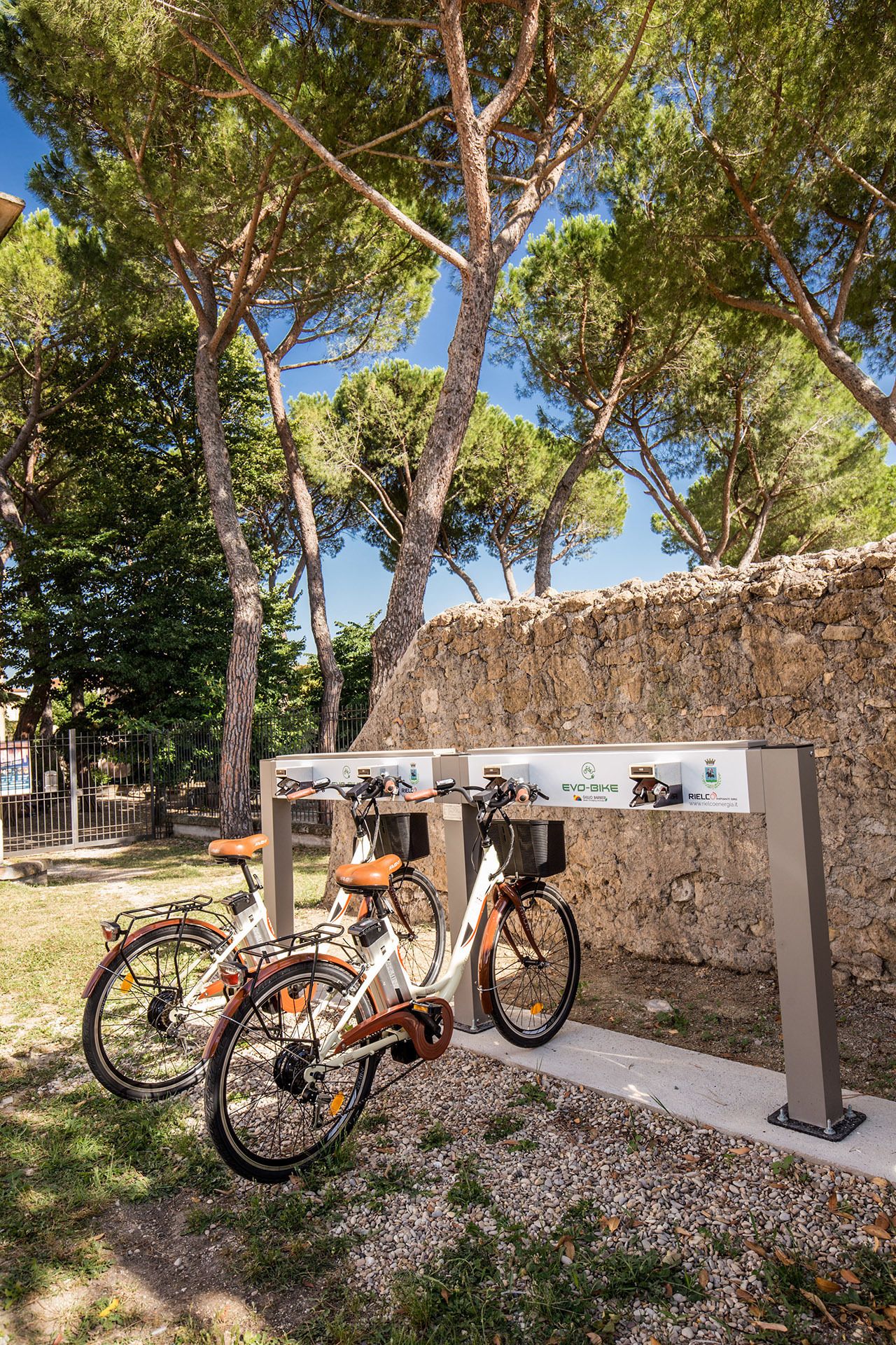 A bike parking area with two bicycles, a stone wall, and trees in the background.