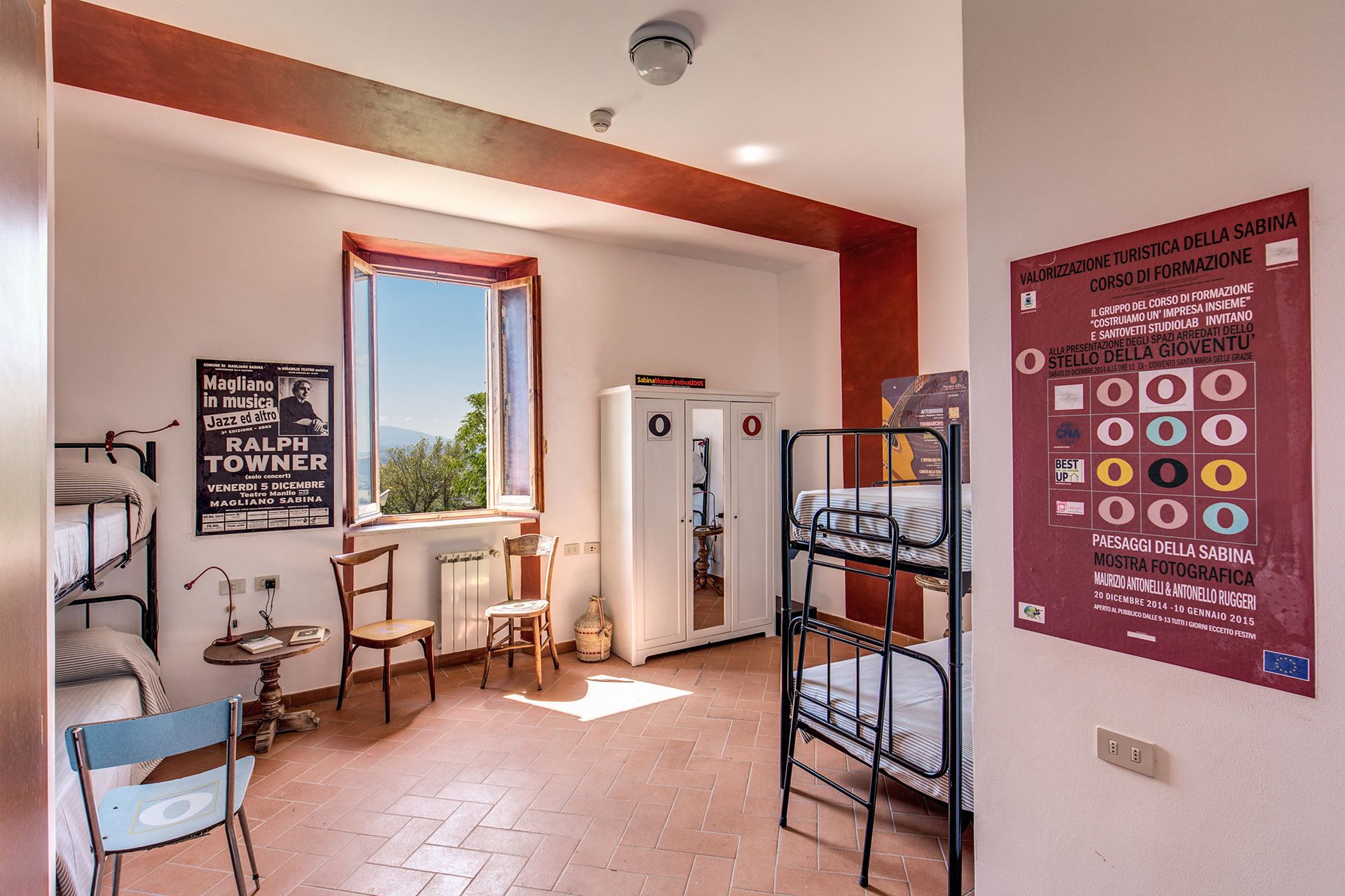 A dormitory room with bunk beds, a radiator, a window with a view, a poster on the wall, and a table with chairs.