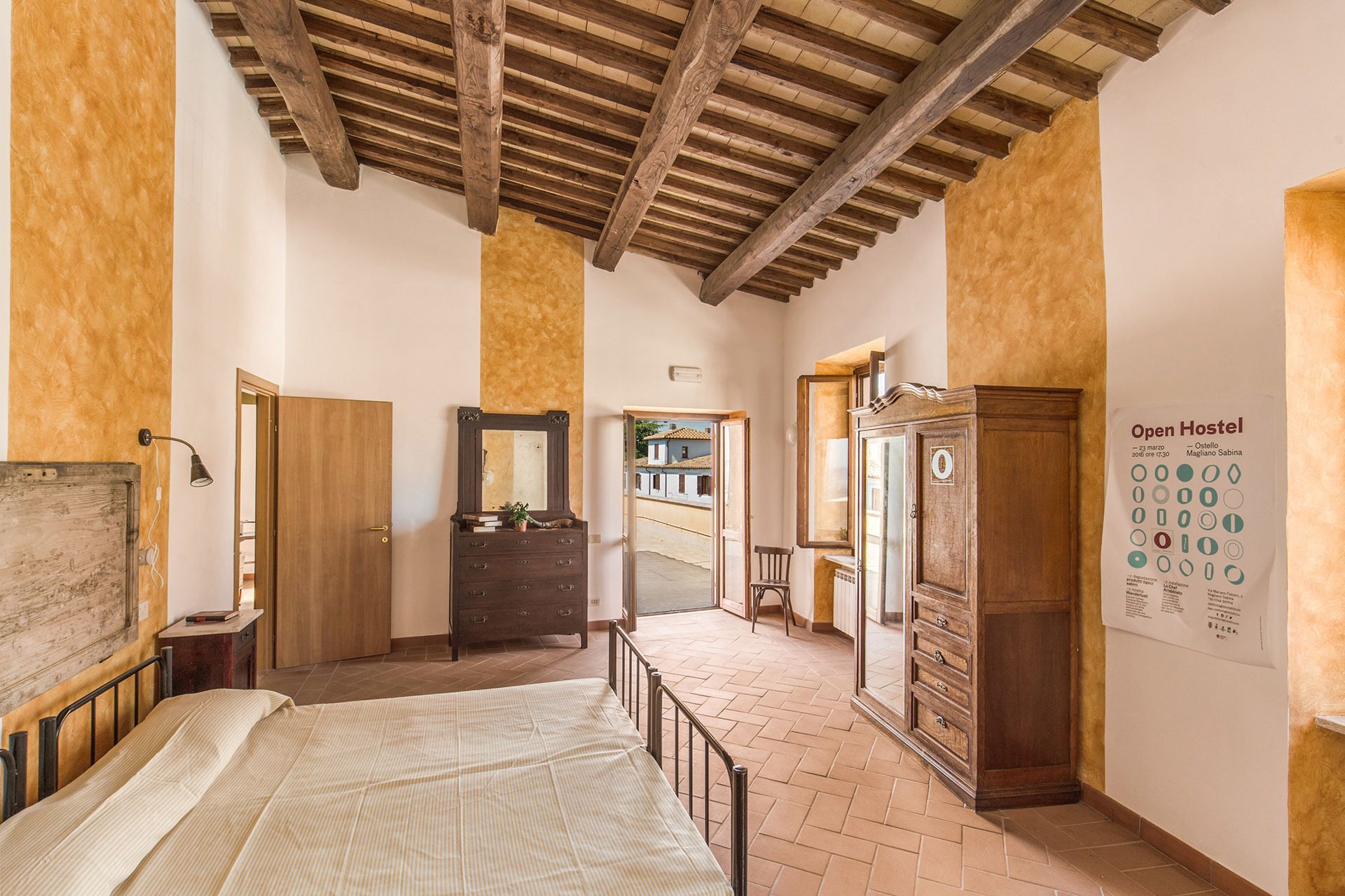 A room with a wooden ceiling, a bed, a dresser, a mirror, and a cabinet. There is a door leading to another room.