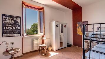 Una habitación con ventana, radiador, armario, una mesa con libros y una cama litera.
