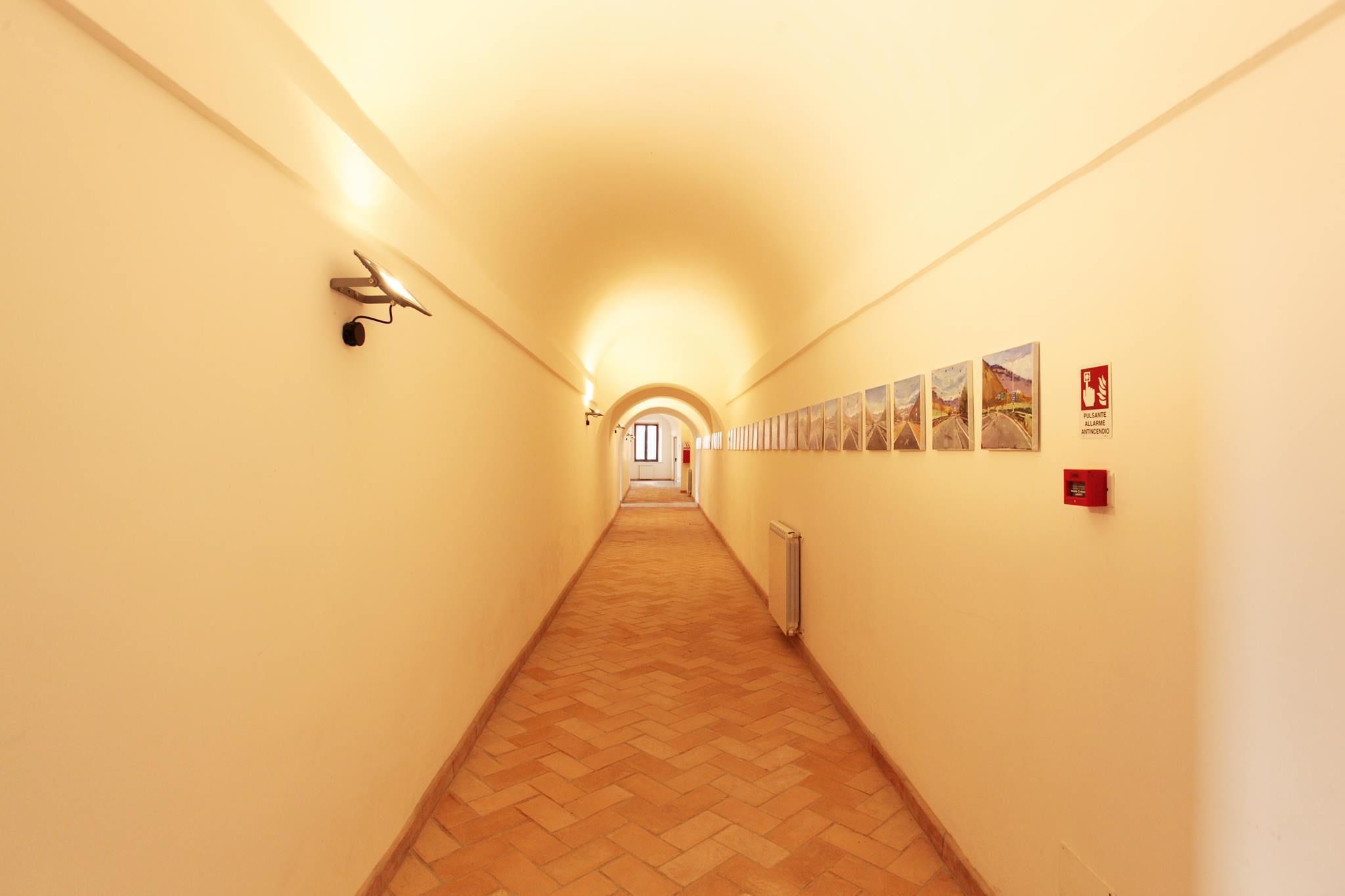 A long, narrow hallway with a wooden floor and light-colored walls. There are several framed pictures on the right wall and a fire alarm on the left.