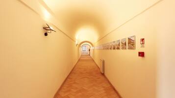 A long, narrow hallway with a wooden floor and light-colored walls. There are several framed pictures on the right wall and a fire alarm on the left.