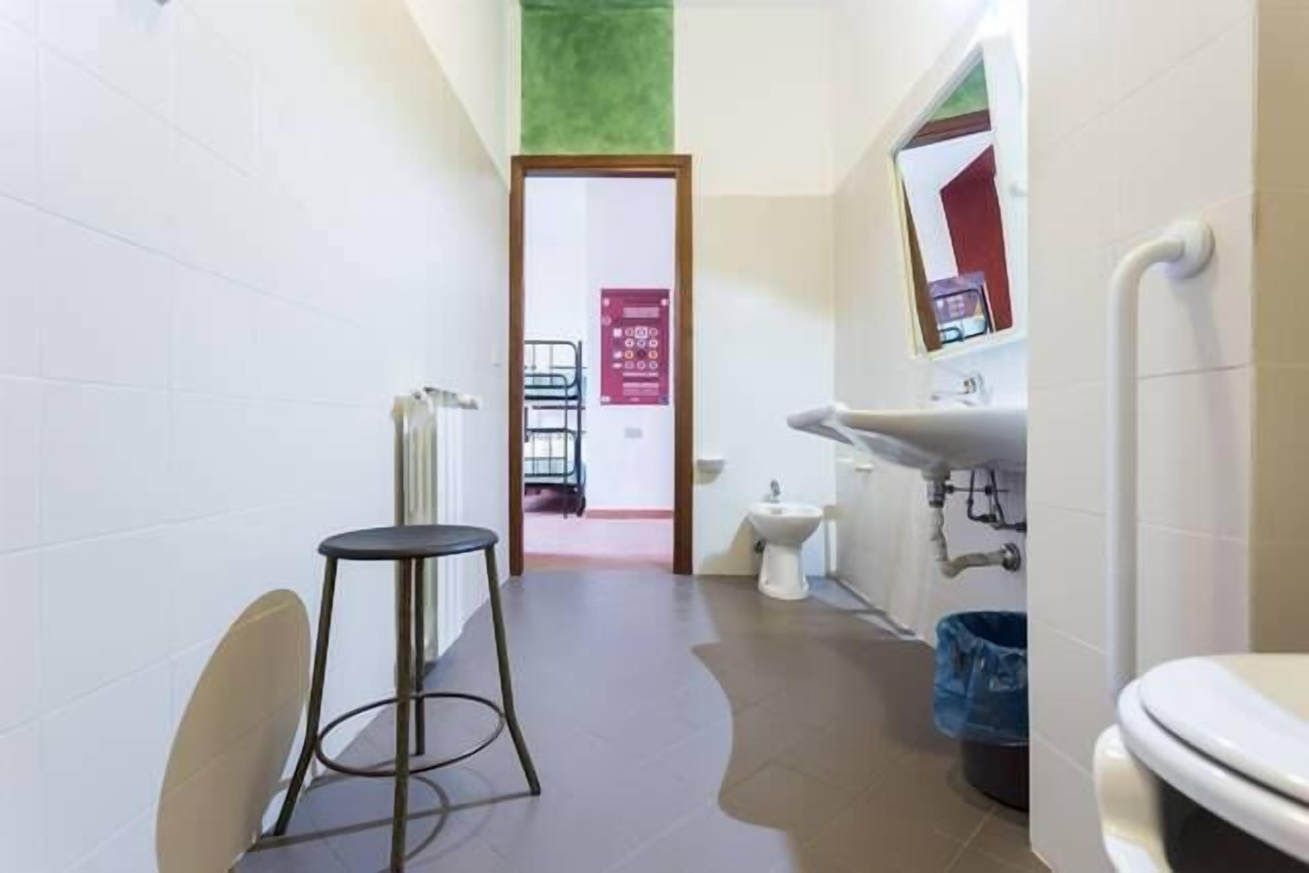 Un pasillo estrecho con un inodoro, lavamanos y una silla. Una puerta da a otra habitación.