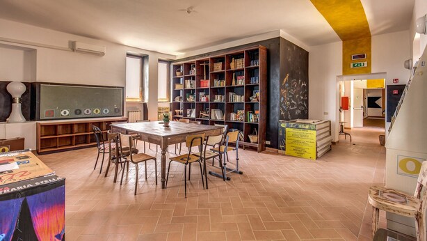 Una biblioteca con un gran escritorio de madera y sillas, rodeada de altas estanterías llenas de libros y objetos decorativos.