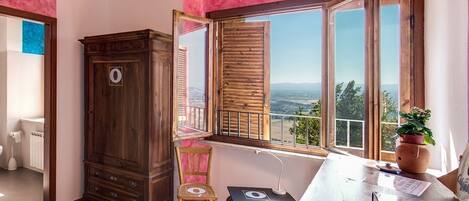 A room with a wooden cabinet, a chair, a table with a laptop, and a window with a view of the landscape.