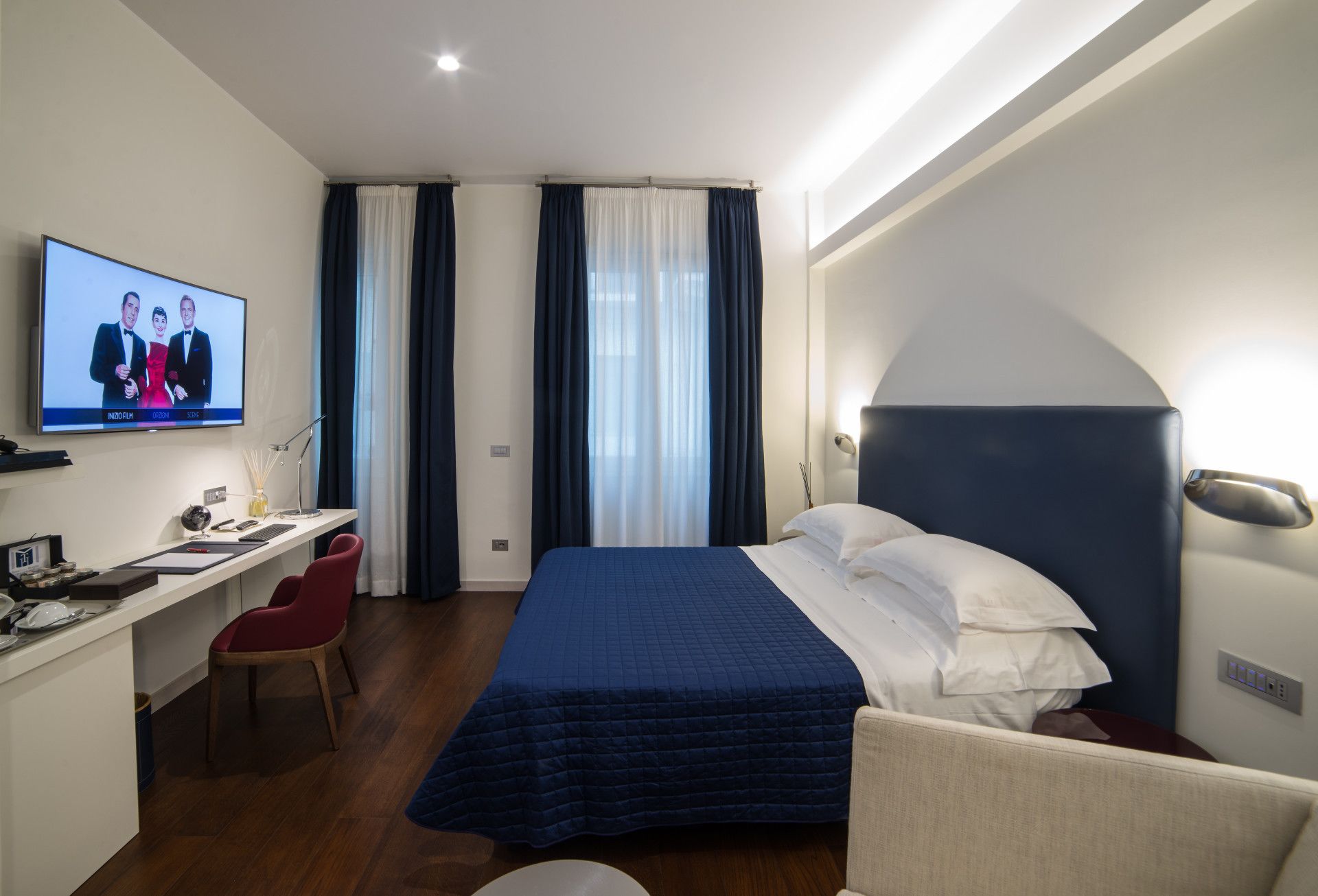 Habitación de hotel con cama, escritorio, silla y televisor mostrando una escena de película.