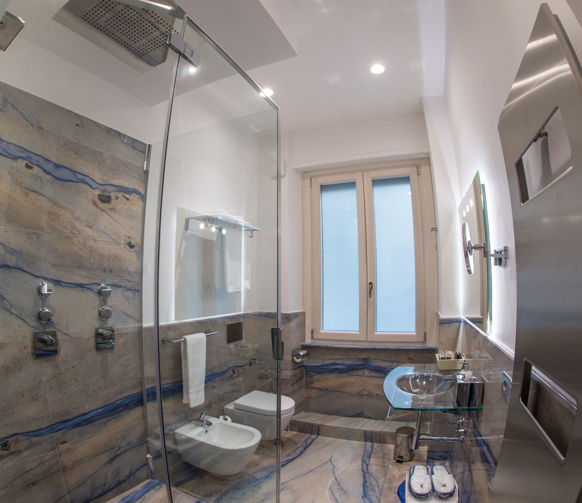 A modern bathroom with a glass-enclosed shower, a marble-tiled wall, a toilet, a sink, and a large window.