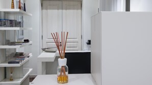 Une pièce moderne et minimaliste avec des murs blancs, une porte blanche et un séparateur de pièce blanc. On y trouve des étagères avec des livres et des objets décoratifs, une table avec une bouteille contenant un liquide et des diffuseurs à bâtonnets, ainsi que trois lampes suspendues.