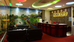 A hotel reception area with a mural of a scenic landscape, a reception desk, and a black sofa. - Minh Kieu Hotel (My Tho)