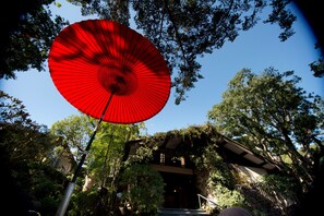 A red umbrella with a metal pole, surrounded by trees and a building with a sloped roof. - Abba Resorts Izu - Zagyosoh (Ito)