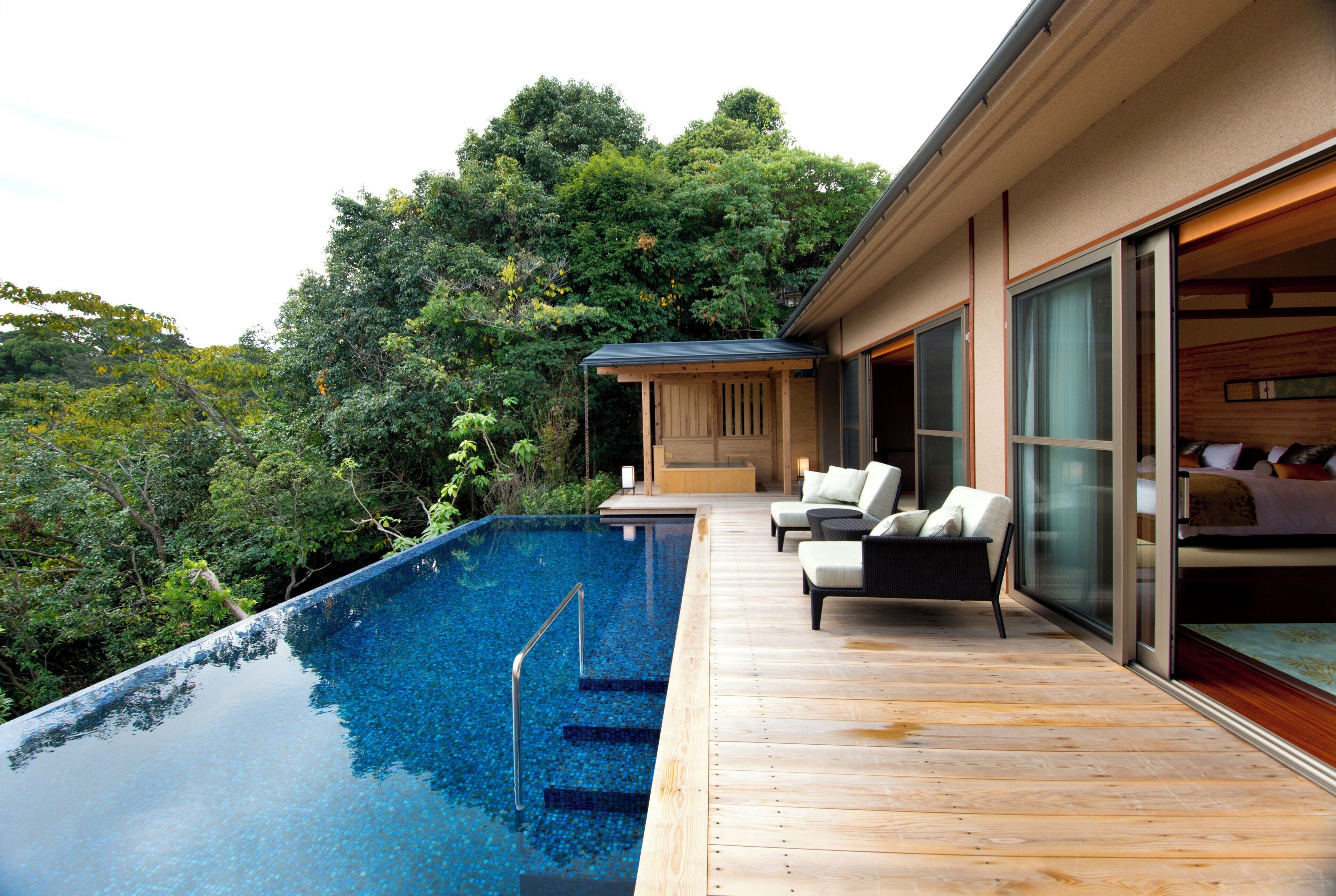 A modern outdoor infinity pool with wooden deck, outdoor seating, and a view of lush greenery.