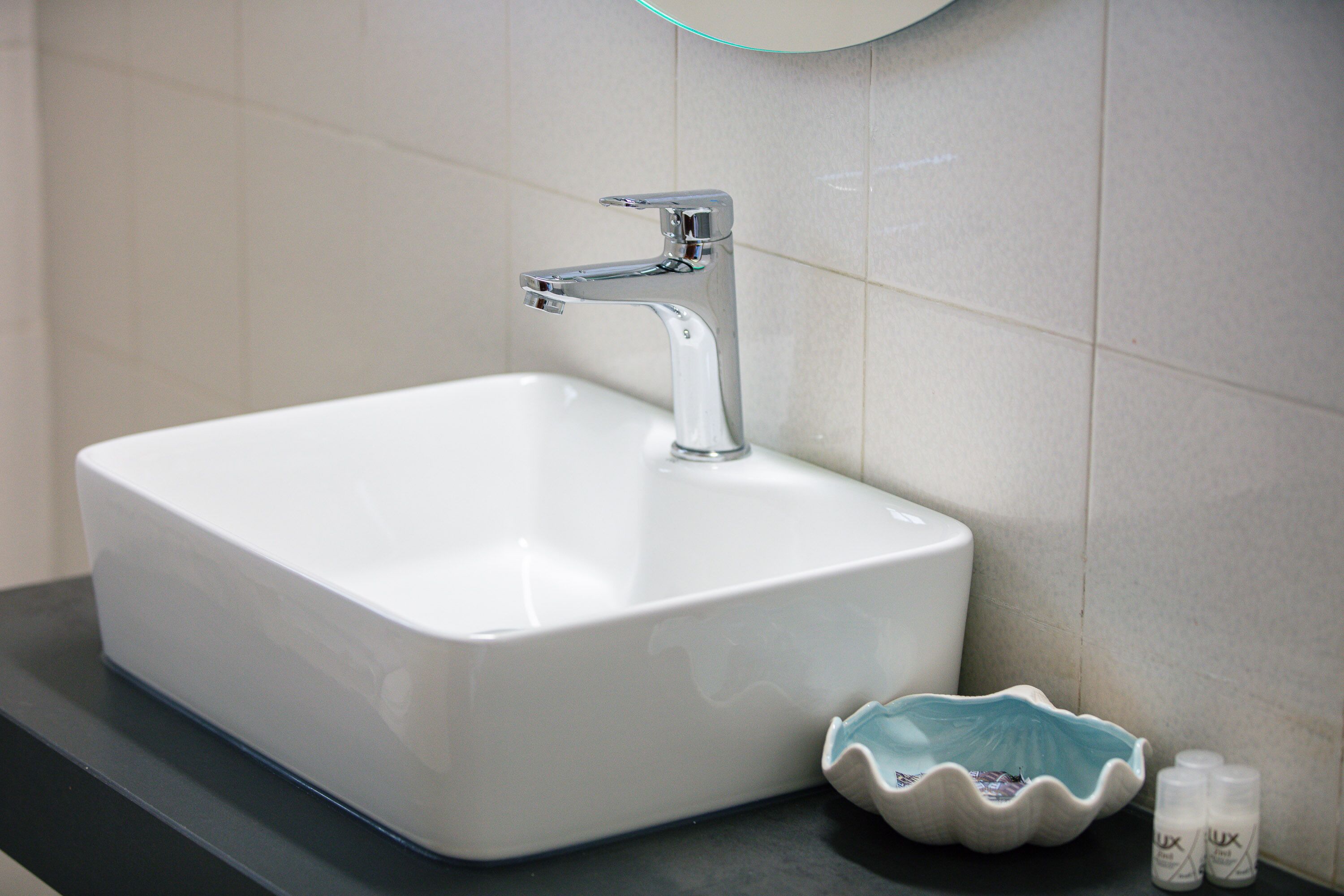 A white rectangular bathroom sink with a chrome faucet, a decorative shell-shaped bowl, and two small bottles of soap on a dark countertop.