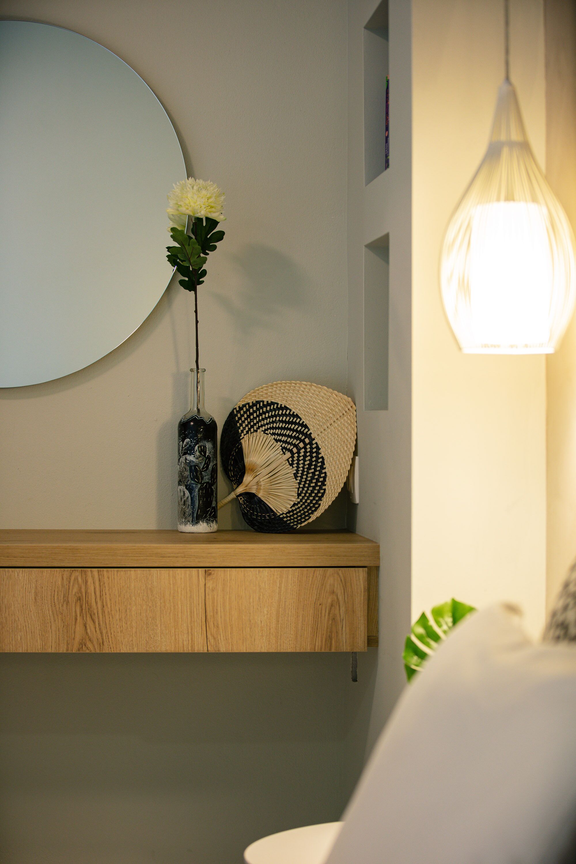 A bathroom with a wooden shelf, a round mirror, a vase with flowers, a woven basket, and a hanging light fixture.