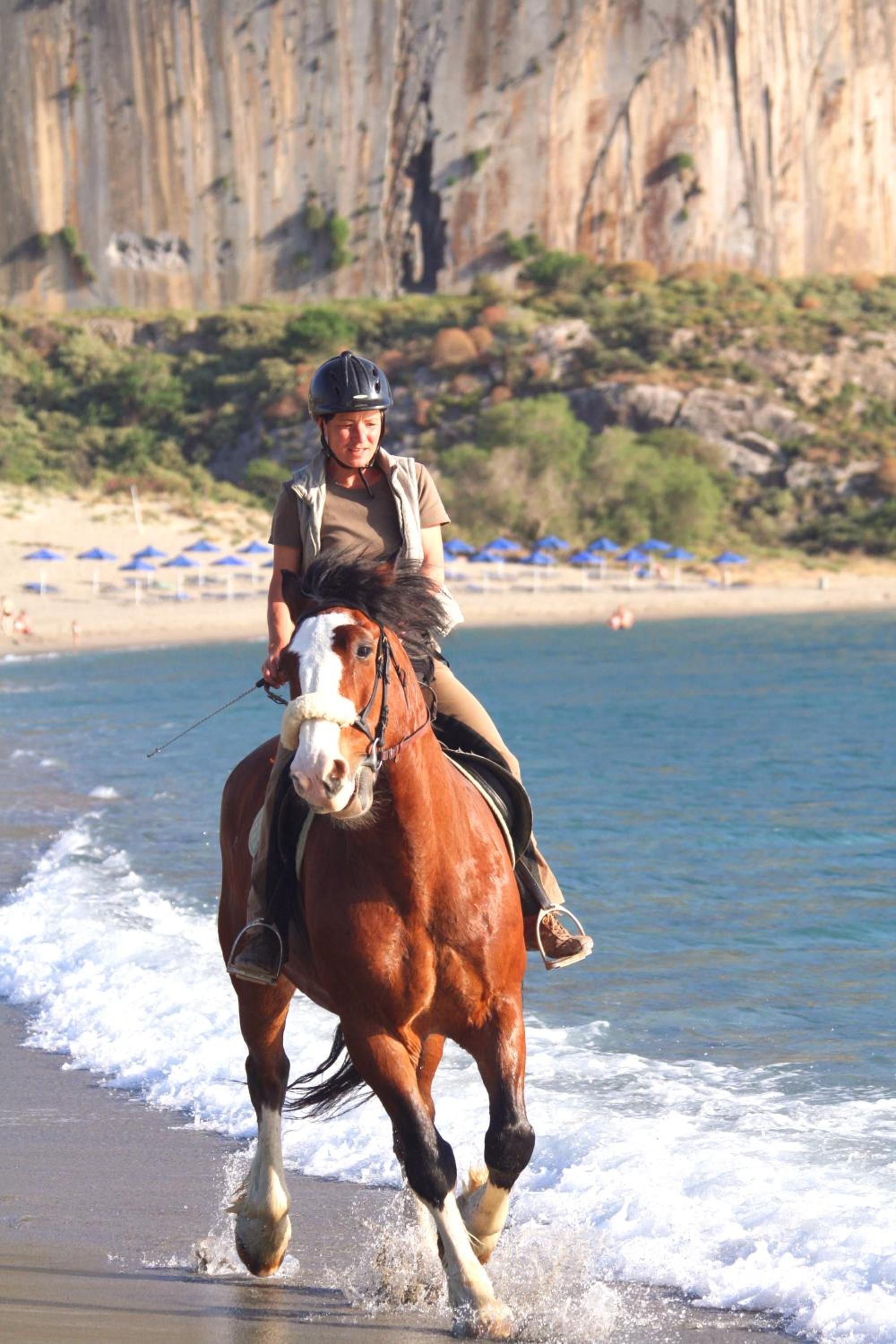 Une personne à cheval, chevauchant le long d’une plage, avec des falaises en arrière-plan.