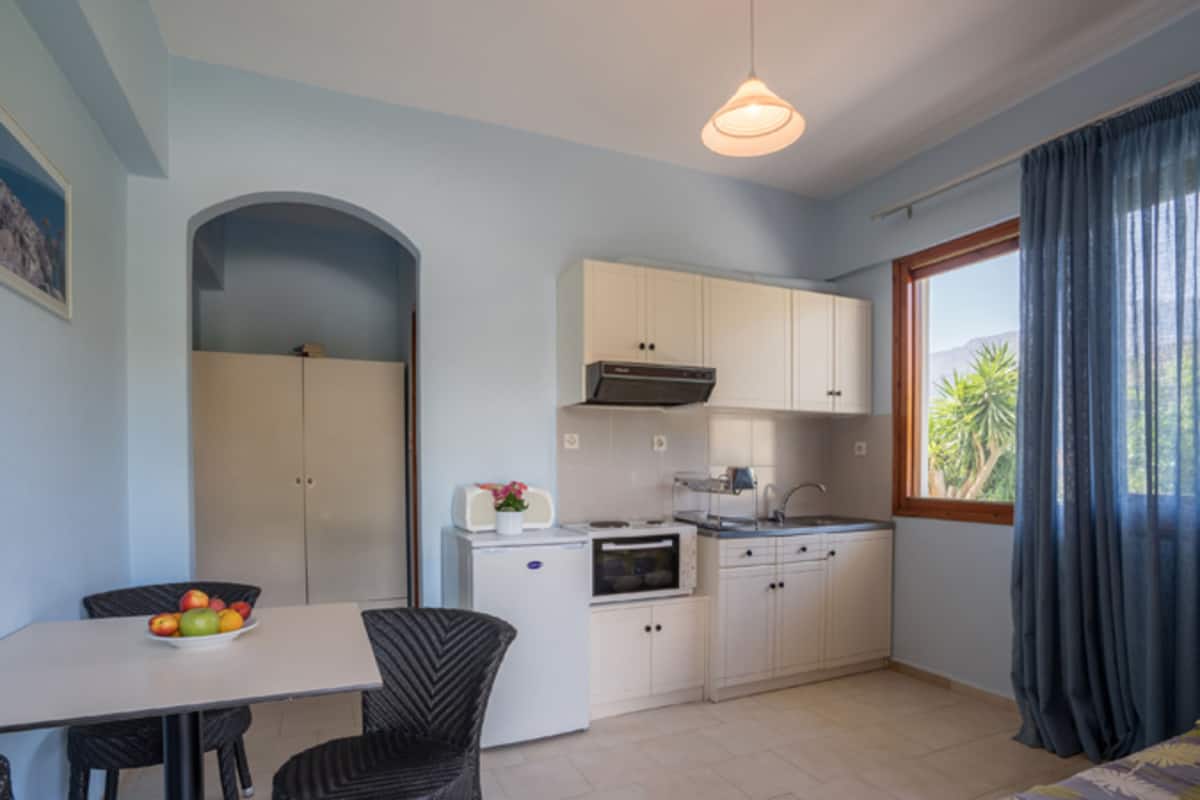 A compact kitchen with white cabinets, a microwave, a toaster, and a sink. There is a dining area with a table and chairs, and a window with curtains.