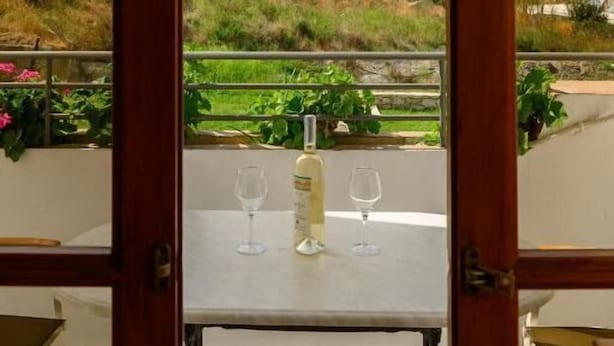 Una mesa con dos copas de vino y una botella, frente a una vista panorámica al aire libre a través de una ventana con marco de madera.