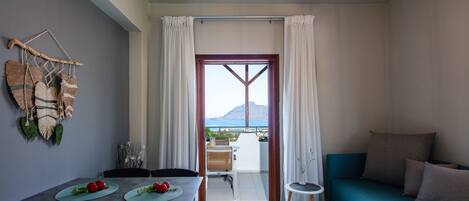 A modern living room with a sofa, a dining table with placemats, and a view of the sea through a large window.