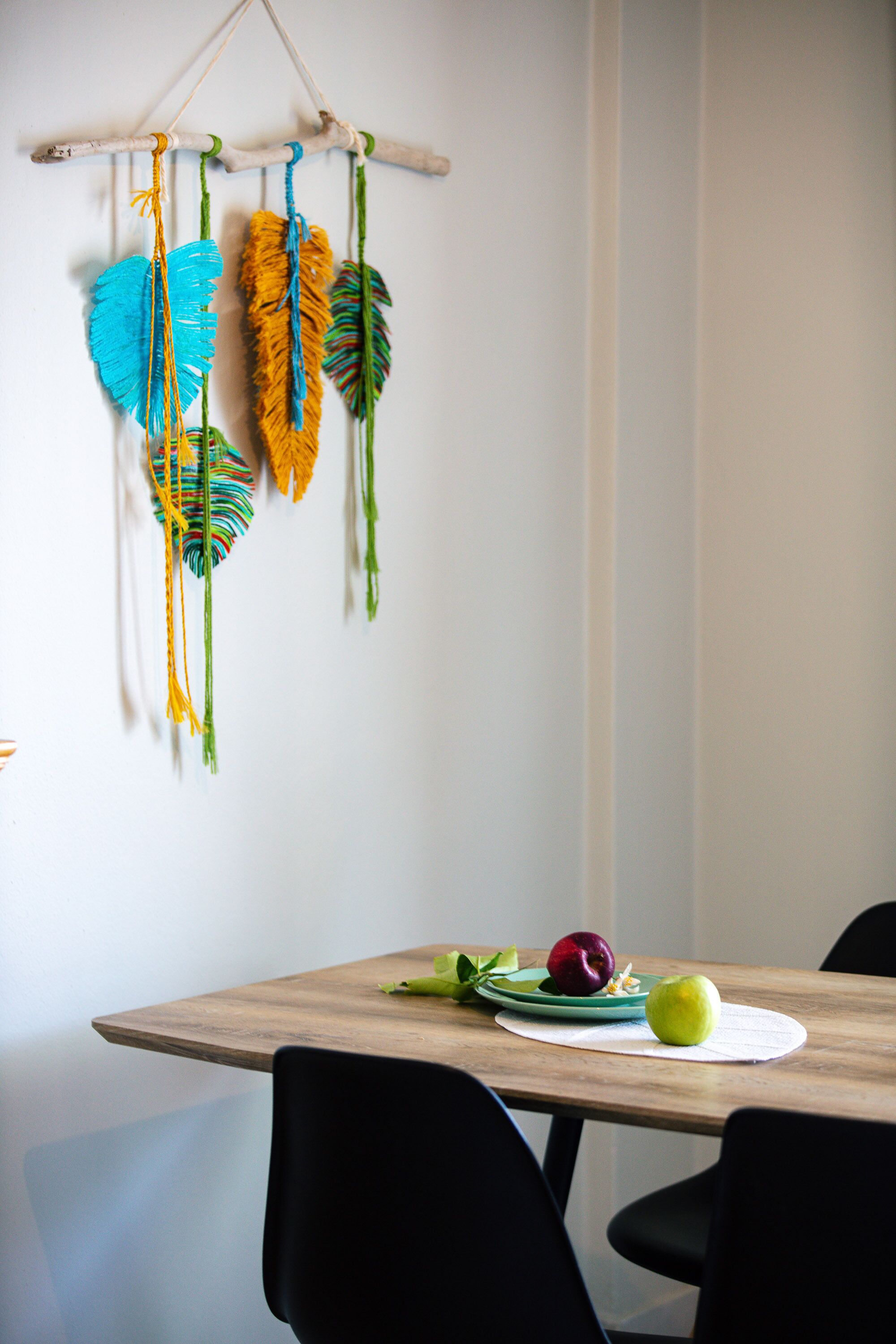 A dining table with a white placemat, a green plate with two apples, and a wooden wall hanging with colorful feathers.
