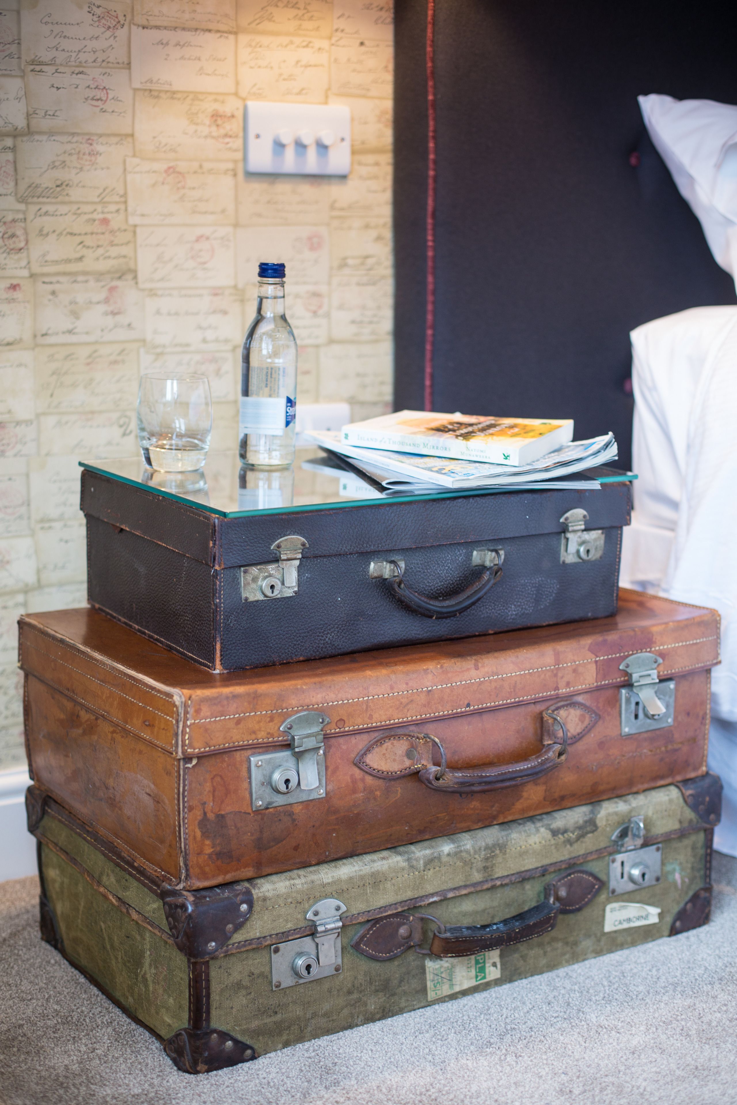 A stack of vintage suitcases with a glass table on top, containing a bottle, glasses, and books.