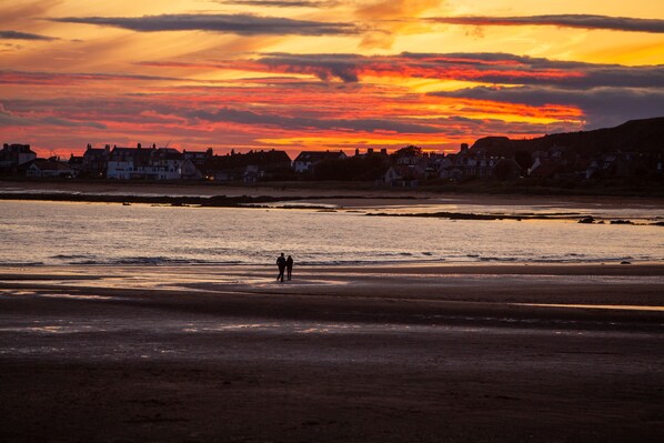 Una playa con dos personas caminando por la orilla, casas al fondo y un atardecer sobre el agua.