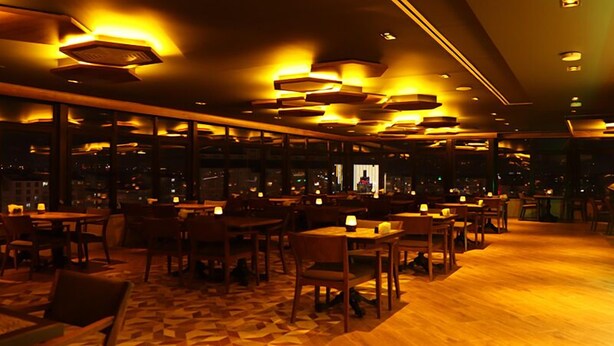 Un restaurante tenuemente iluminado con varias mesas y sillas, ventanas amplias y una alfombra estampada.