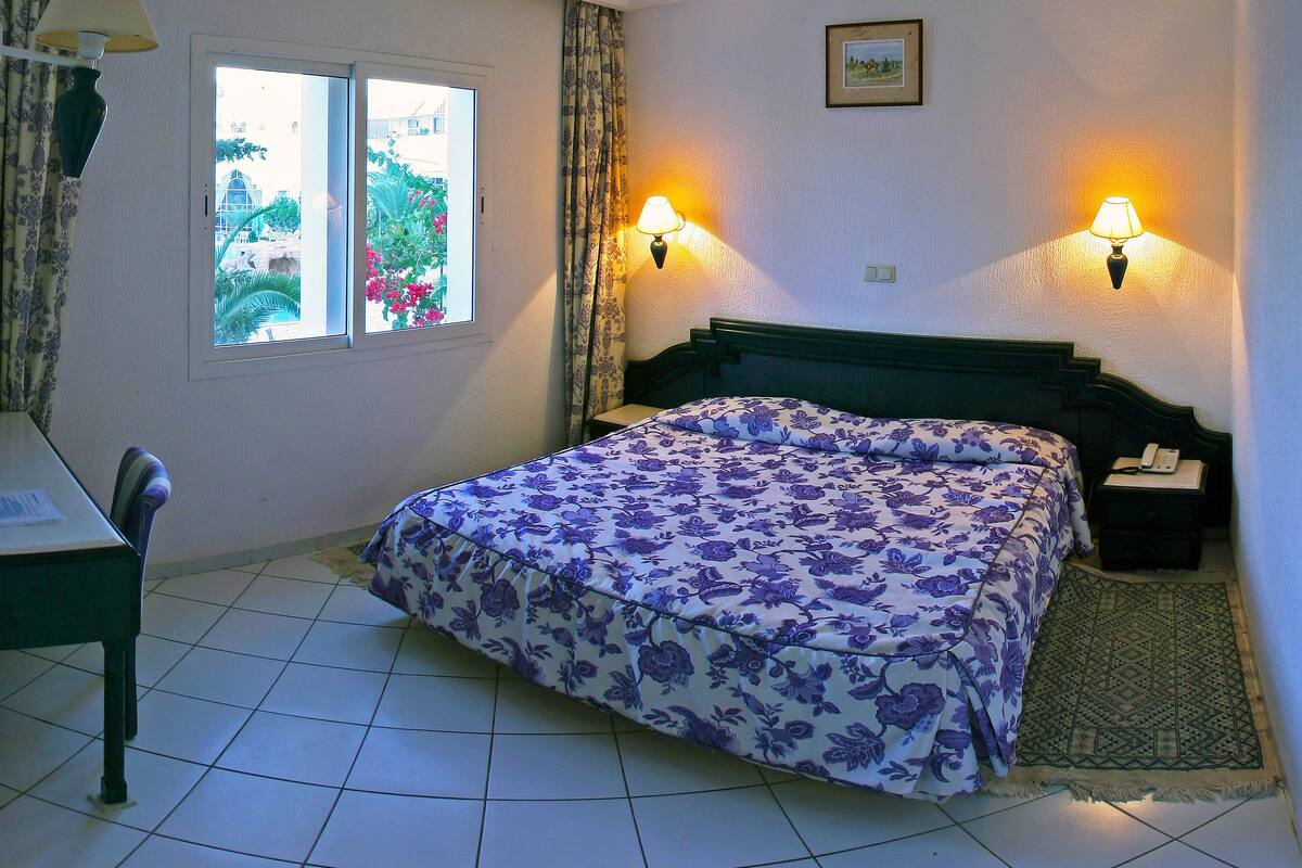 Una habitación de hotel con cama, dos lámparas de noche, ventana con cortinas, una silla y una mesita.