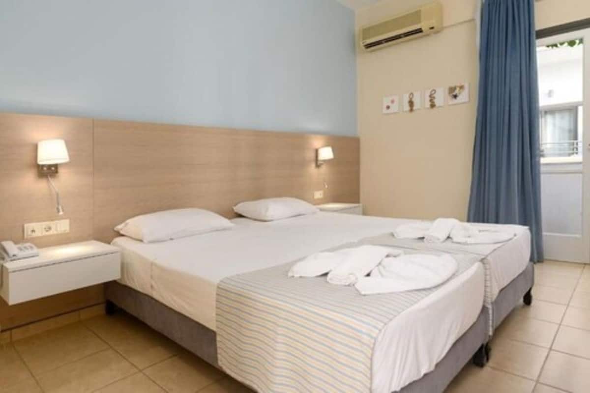 A hotel room with a bed, bedside tables, a phone, an air conditioning unit, and a window with curtains.