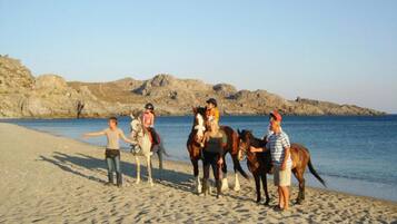 A beach with people and horses, rocky cliffs in the background, and clear skies.