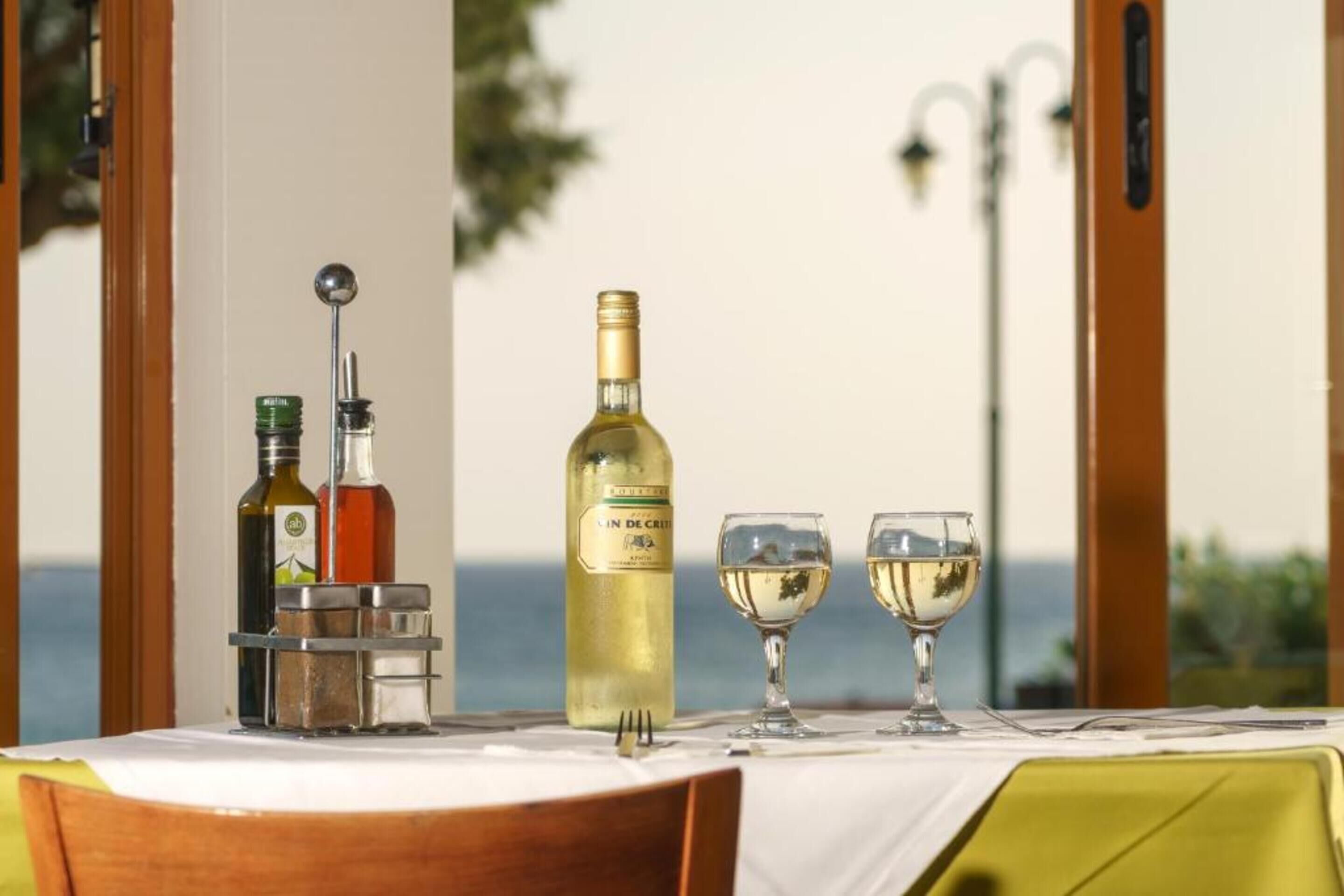 A table set with a bottle of white wine, two glasses, and a bottle of olive oil, with a view of the sea in the background.