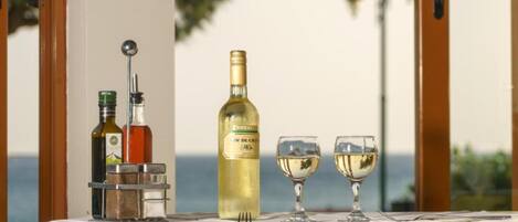 Una mesa dispuesta con una botella de vino blanco, dos copas y una botella de aceite de oliva, con vista al mar de fondo.