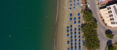 Vista aérea de una playa con hileras de sombrillas azules, un estacionamiento y edificios al fondo.