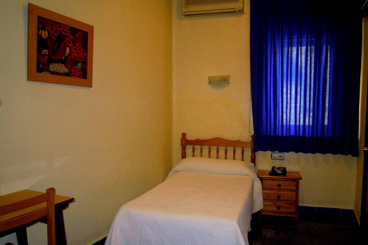 Un dormitorio pequeño con una cama individual, una mesita de noche de madera, una silla, un aire acondicionado instalado en la pared y una ventana con cortinas azules.
