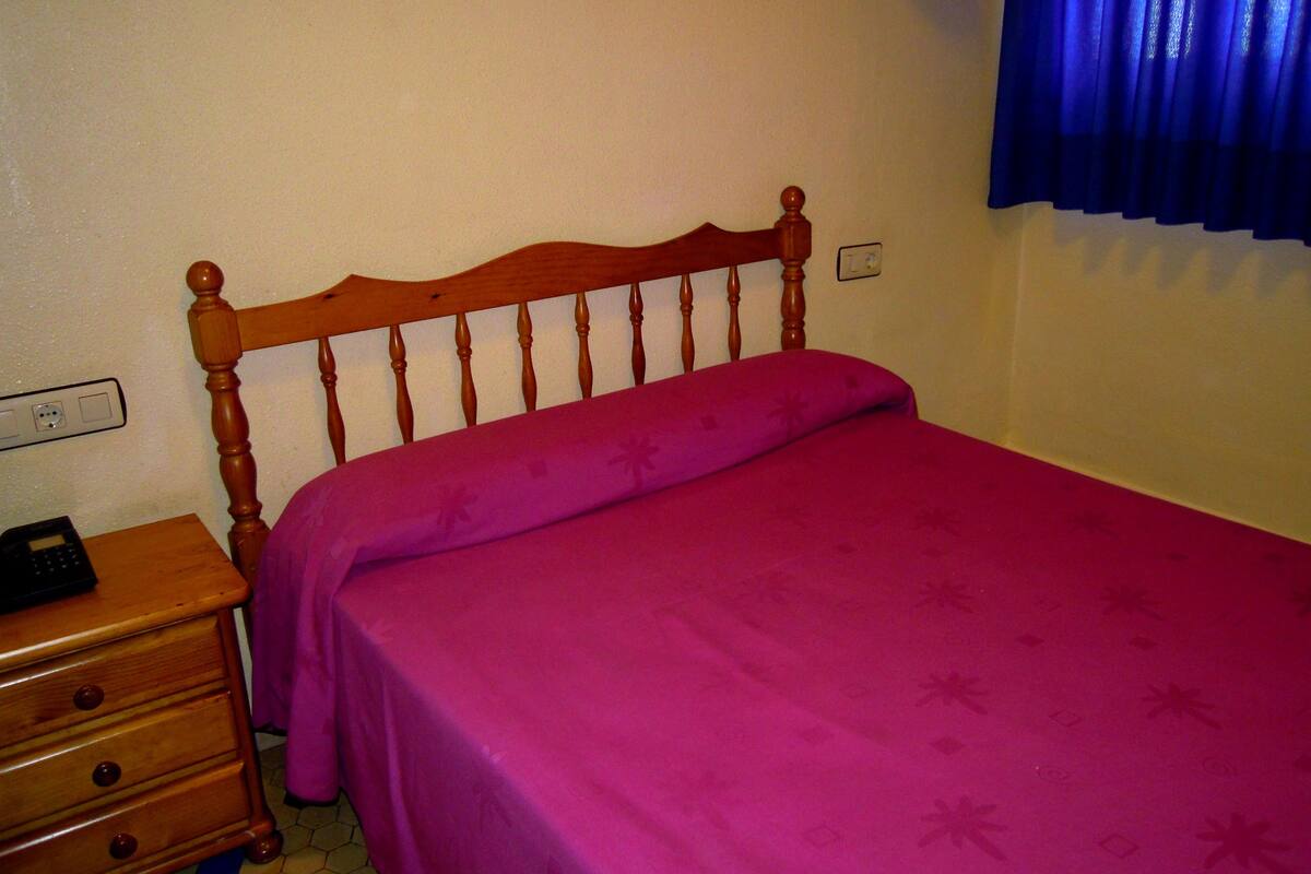 Un dormitorio con cabecera de madera, colcha rosa, mesita de noche de madera y cortina azul.