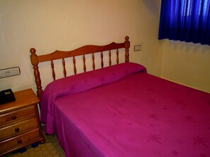 A bedroom with a wooden headboard, a pink bedspread, a wooden nightstand, and a blue curtain. - Hotel Europa (Valencia)