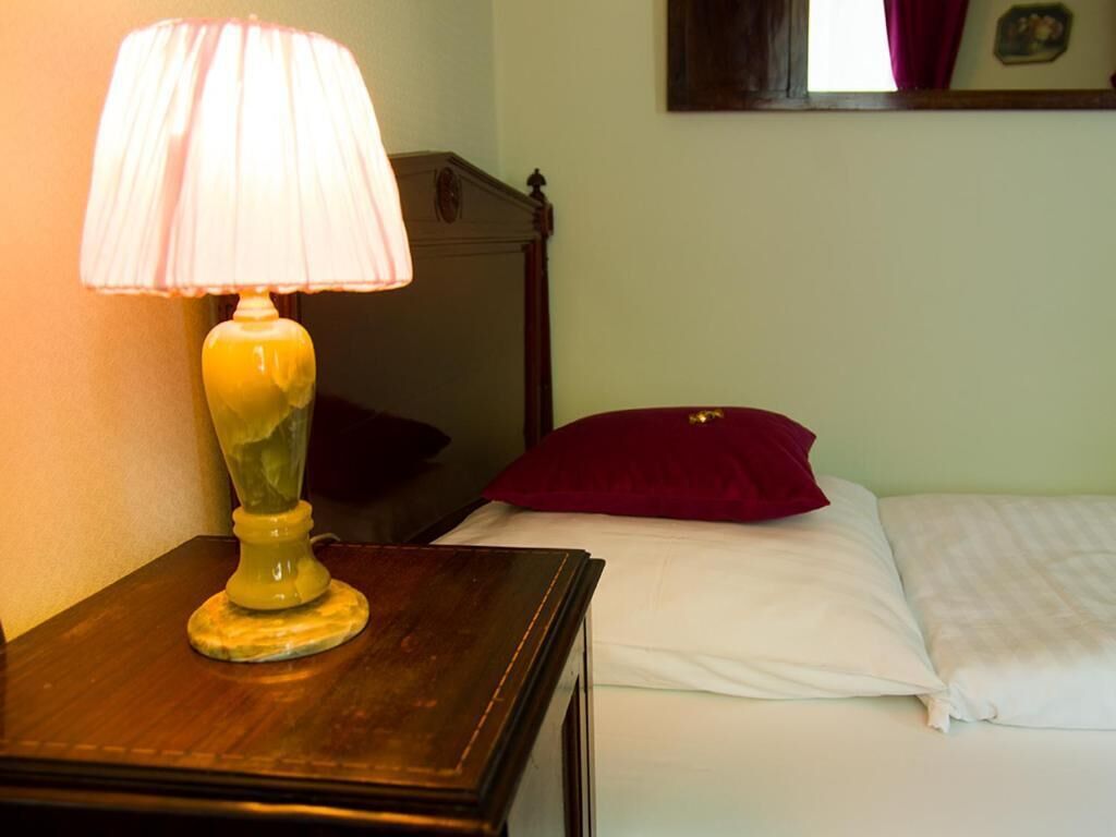 A wooden nightstand with a lamp, a bed with white bedding and a red pillow, and a wooden headboard.