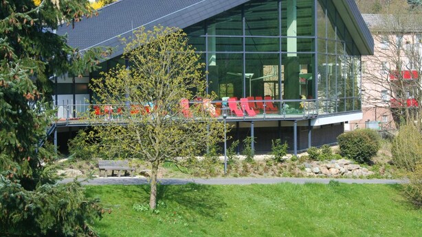 A modern building with large glass windows, a sloped roof, and a small garden area with a bench. - Hotel Victoria (Schlangenbad)