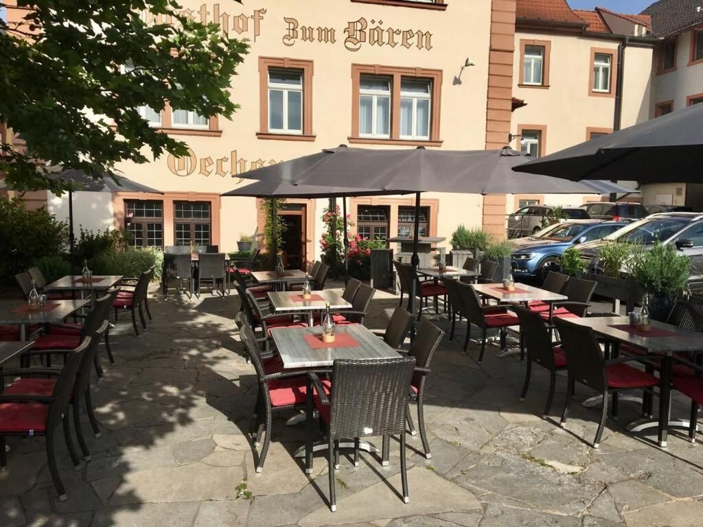 An outdoor cafe area with tables and chairs, umbrellas, and a building with a sign in the background.
