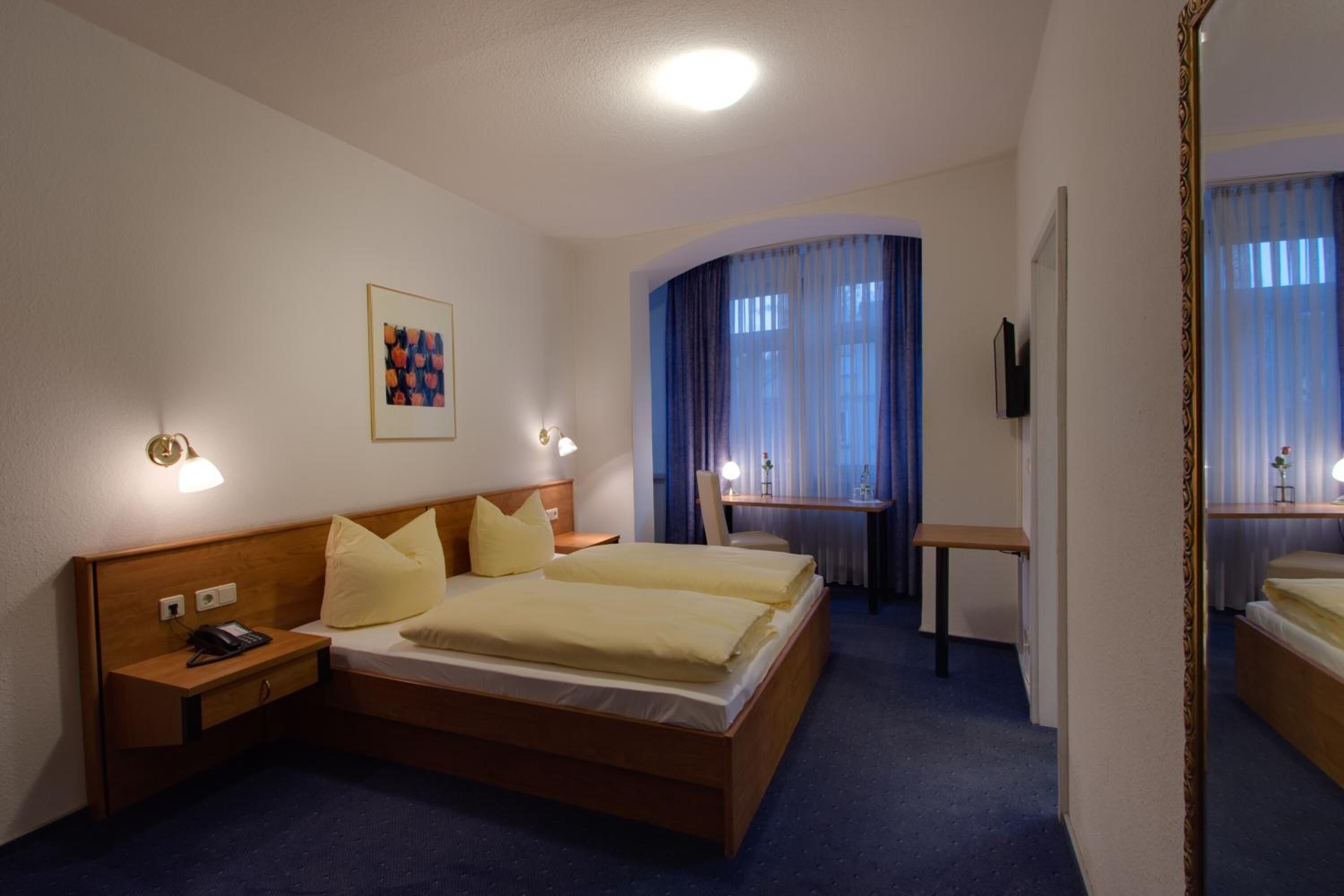 Ein Hotelzimmer mit Doppelbett, Nachttischen, einem Schreibtisch, einem Sessel und einem Spiegel, sowie einem Bild an der Wand.