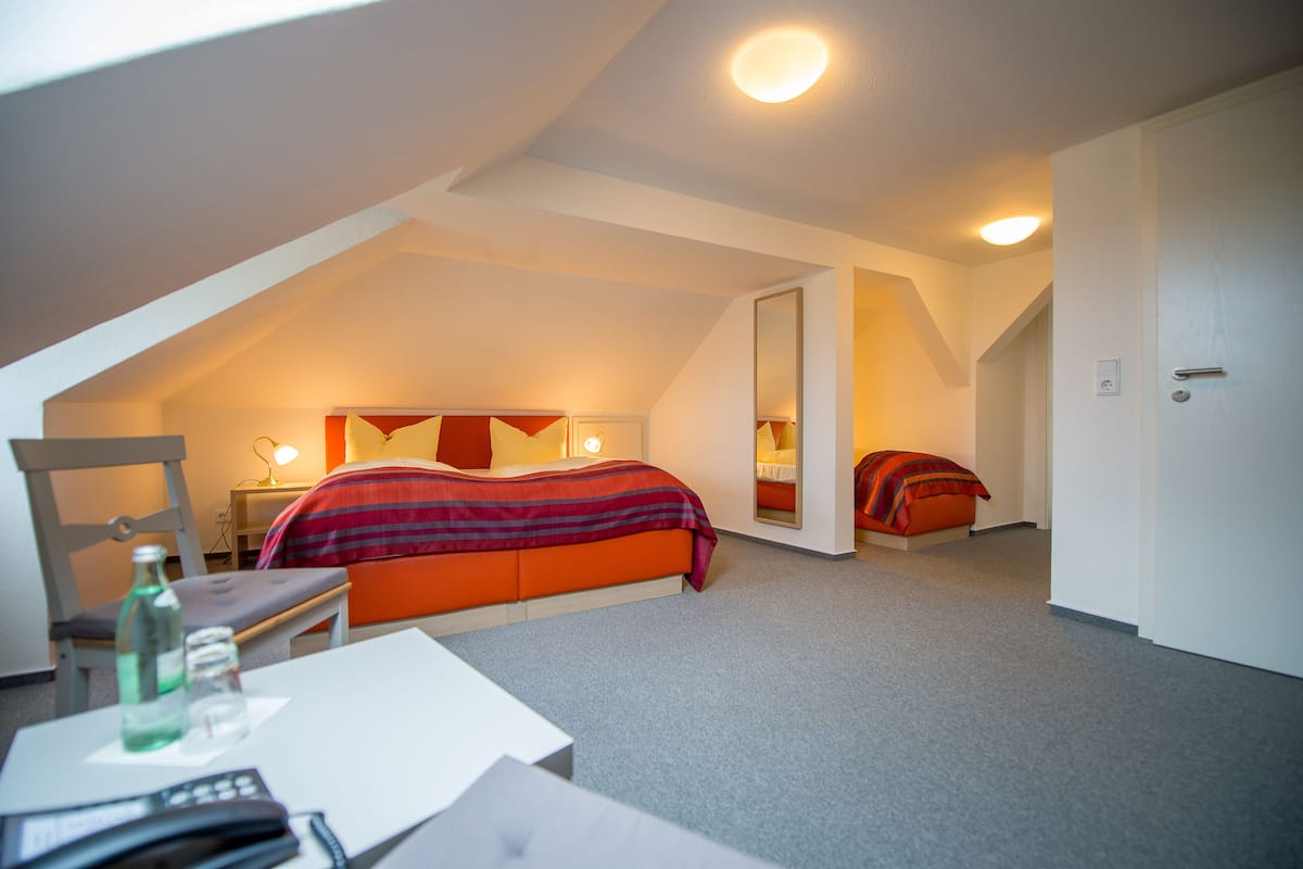 A bedroom with a sloped ceiling, a bed with orange bedding, a bedside table with a lamp, and a chair.