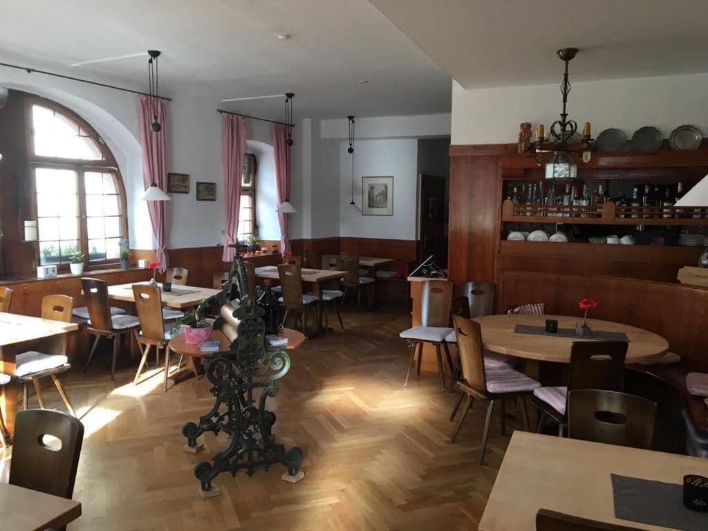 A restaurant with wooden flooring, multiple tables and chairs, a bar area with shelves, and a large window with curtains.