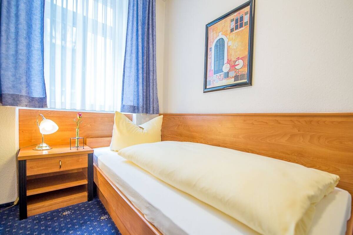 A hotel room with a wooden headboard, a single bed, a nightstand with a lamp, and a framed picture on the wall.