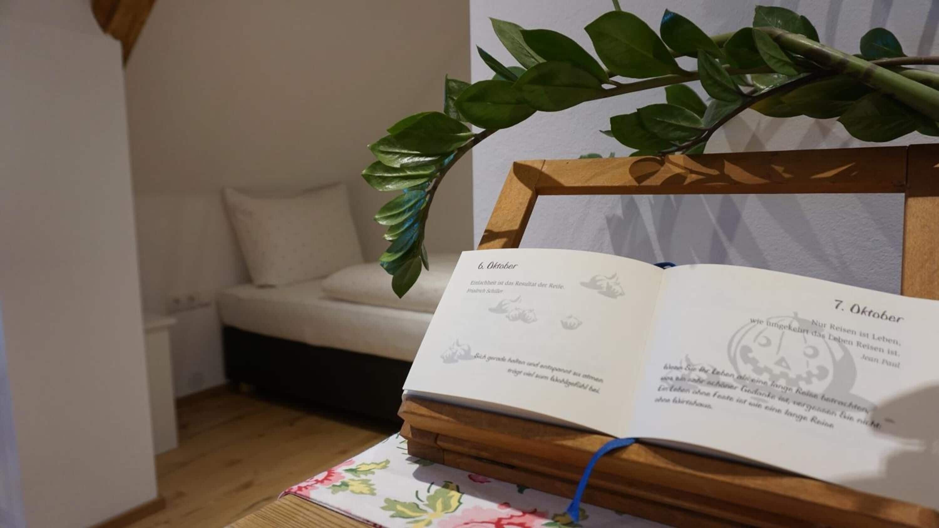 A room with a bed, a plant, and an open book on a wooden stand.