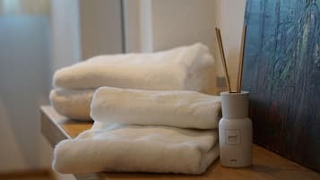 Des serviettes blanches, soigneusement pliées, sont empilées sur une surface en bois, à côté d’un diffuseur blanc muni de bâtonnets en roseau.