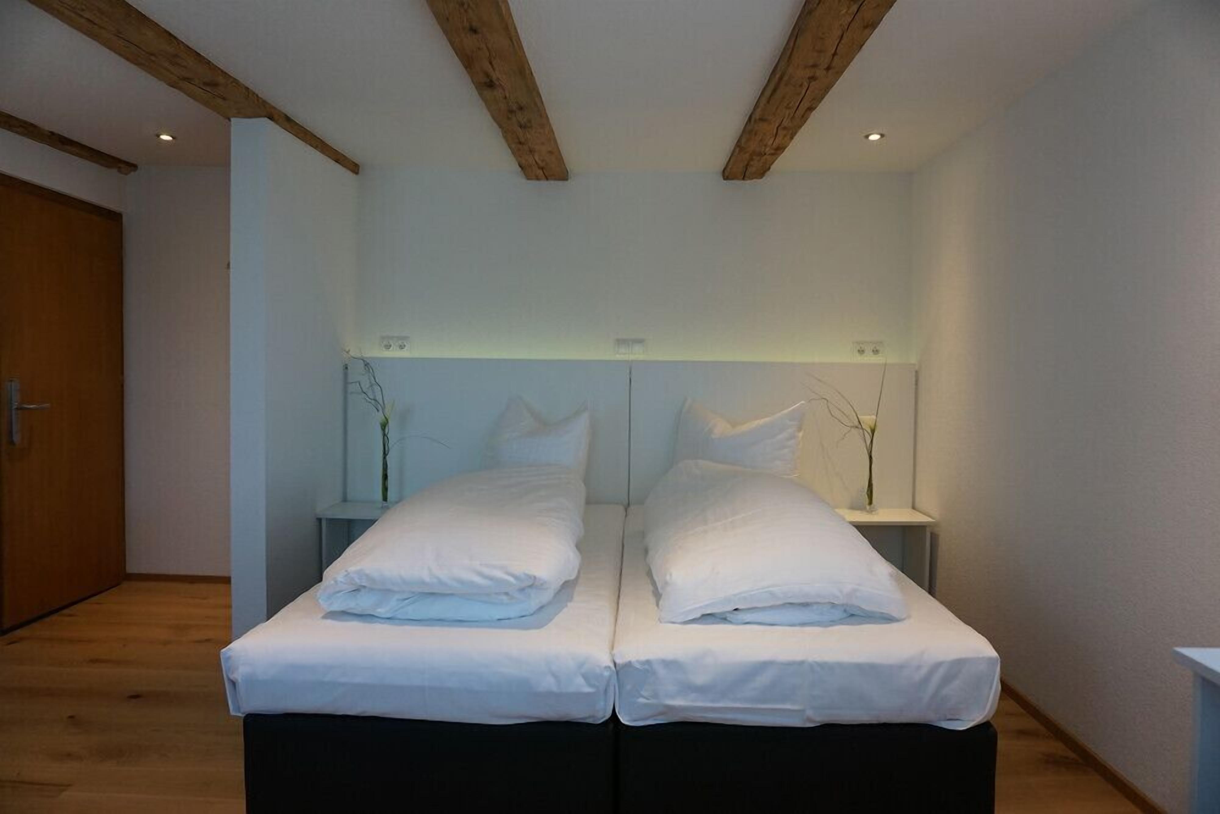 Un lit double avec du linge de lit blanc, dans une chambre dont le plafond est orné de poutres en bois, et une petite table de chevet sur laquelle est posé un vase.