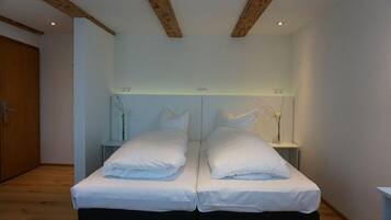 Un lit double avec du linge de lit blanc, dans une chambre dont le plafond est orné de poutres en bois, et une petite table de chevet sur laquelle est posé un vase.