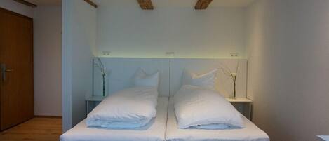 Una cama doble con sábanas blancas en una habitación con vigas de madera en el techo y una mesita de noche con un florero.
