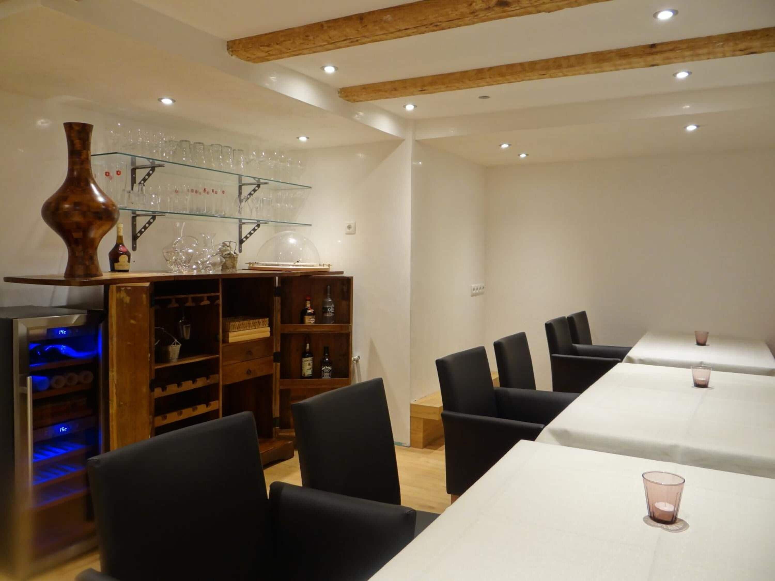 Un comedor con una mesa larga, sillas negras, un estante de madera con cristalería y un jarrón decorativo.