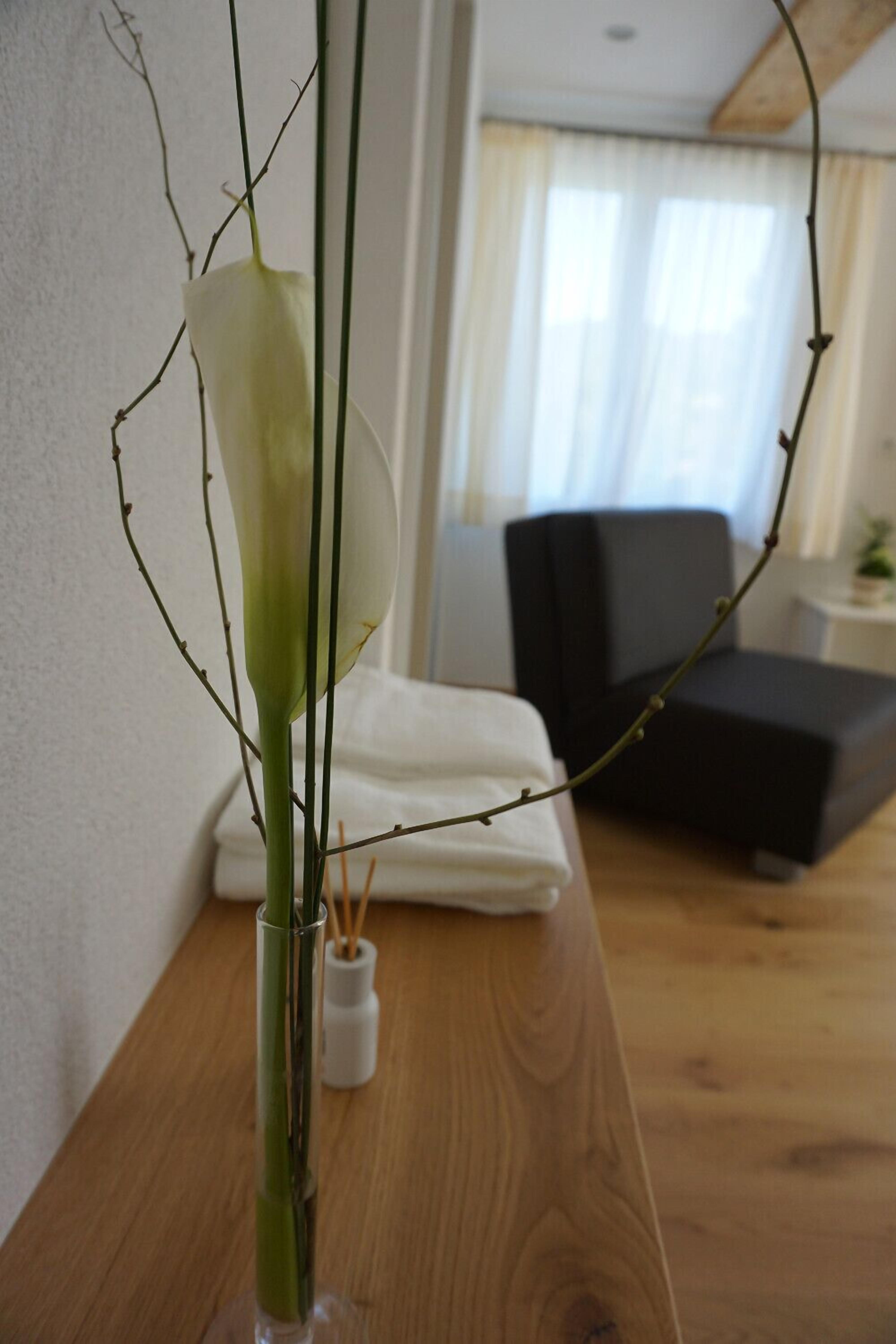 Et værelse med et træbord, en hvid vase med en plante, en sort stol og en seng med hvide sengetæpper.