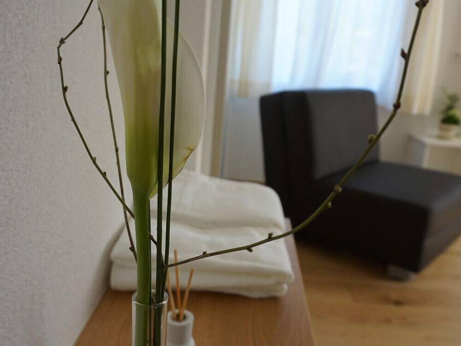 Una habitació amb una taula de fusta, un vas blanc amb una planta, una cadira negra i un llit amb roba de llit blanca.