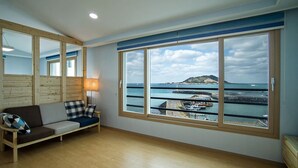 A room with a large window overlooking a coastal area, a wooden floor, and a sofa with cushions. - Sleep in the Sea (Jeju City)